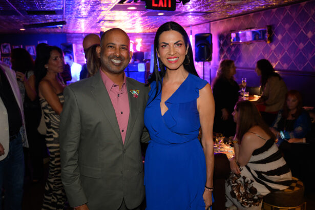 A smiling man in a suit and a woman in a blue dress stand together at a lively indoor event, surrounded by people socializing in a warmly lit room. Lifestyle