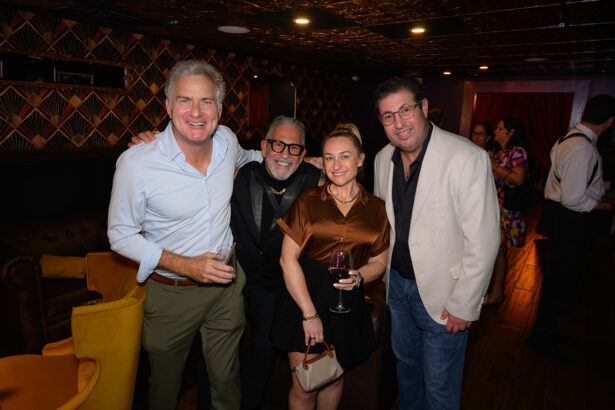 Four adults pose together at a lively indoor event. Three men and one woman smile at the camera, holding drinks, with other guests mingling in the warmly lit, stylish room in the background. Lifestyle