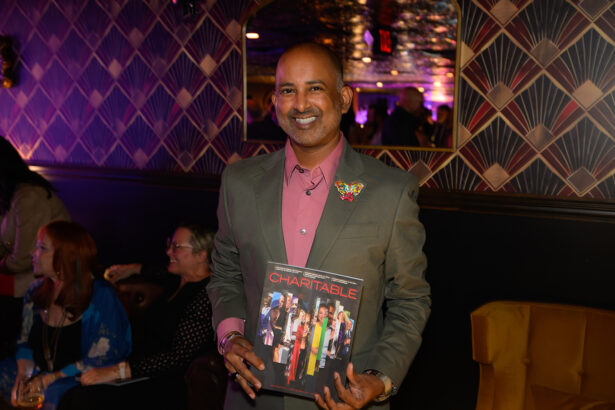 A smiling man in a suit holds a book titled “CHARITABLE” at an indoor event with people seated and colorful, art deco wall patterns in the background. Lifestyle