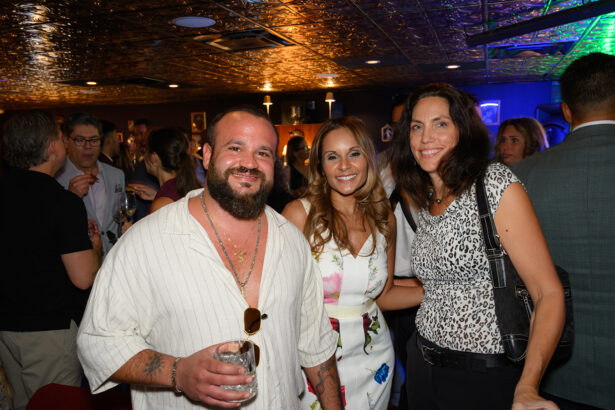 Three people smile at the camera at a crowded indoor event. The man on the left holds a drink, while the two women stand close together. The background shows other guests socializing under warm lighting. Lifestyle
