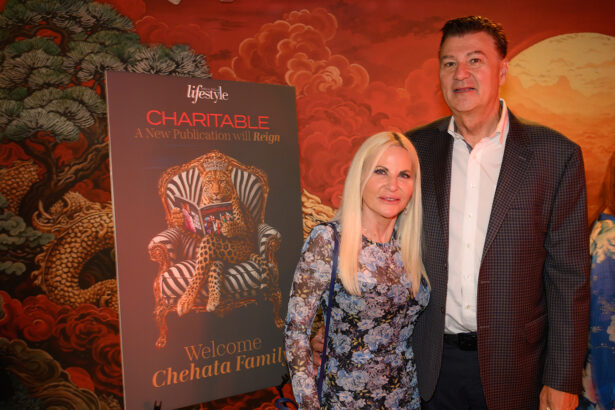 A woman and man stand beside a poster that reads “CHARITABLE: A New Publication will Reign. Welcome Chehata Family.” The background features a decorative dragon mural and warm lighting. Lifestyle
