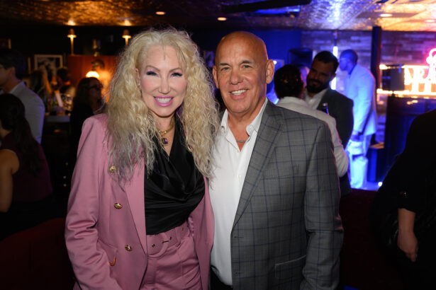 Two people stand and smile at a dimly lit indoor event. The woman wears a pink suit and has curly blonde hair; the man wears a gray checkered jacket. Other people mingle in the background. Lifestyle