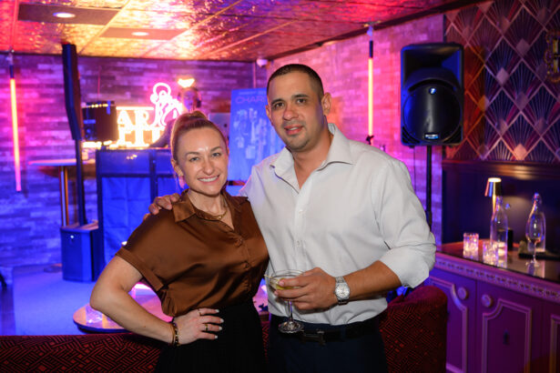 A smiling woman and man pose together at a brightly lit indoor event, with pink and purple neon lights and a stage in the background. The man holds a drink. Lifestyle