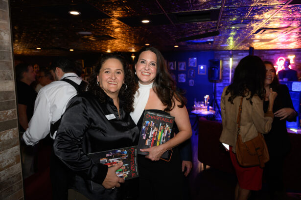 Two women at an indoor event smile and hold magazines titled “CHARITABLE.” The background shows other people mingling, dim lighting, and colorful lights. Lifestyle
