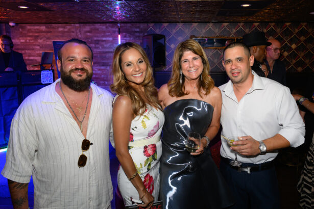 Four adults dressed in semi-formal attire smile and pose together at an indoor party, holding drinks. The background features a DJ booth, colorful lights, and patterned walls. Lifestyle