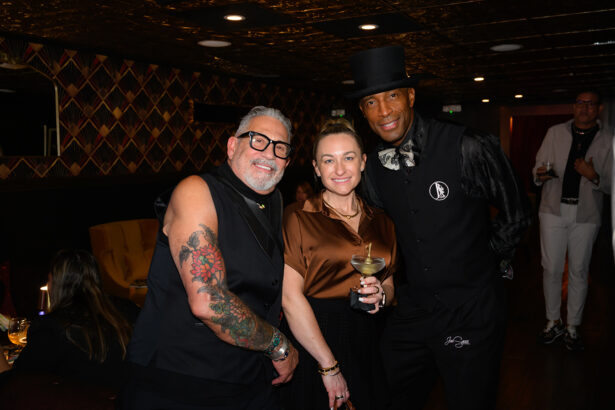 Three people pose together at a stylish indoor event. One man has tattooed arms and glasses, the woman in the center holds a cocktail, and the other man wears a black suit with a top hat. The background is dimly lit and elegant. Lifestyle