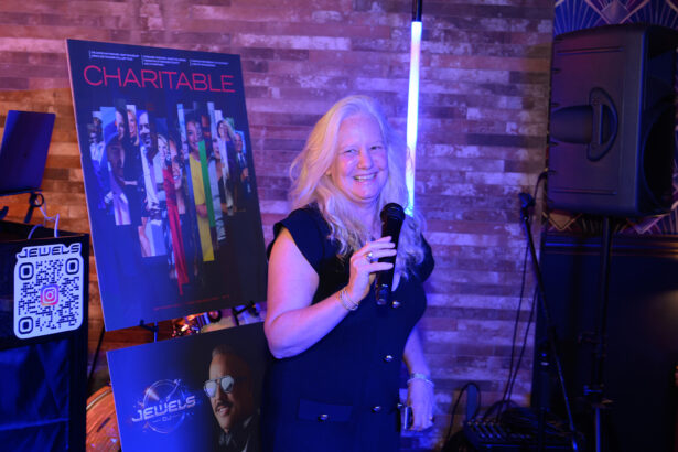 A woman with long white hair holds a microphone and smiles, standing next to posters for “Charitable” and “Jewels.” The background features a brick wall and audio equipment. Lifestyle
