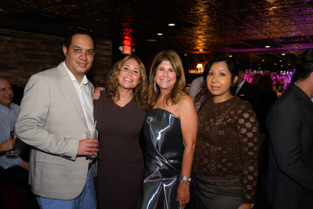 Four adults pose and smile together at a dimly lit indoor event, with a bar and other people visible in the background. The group is dressed in semi-formal attire. Lifestyle
