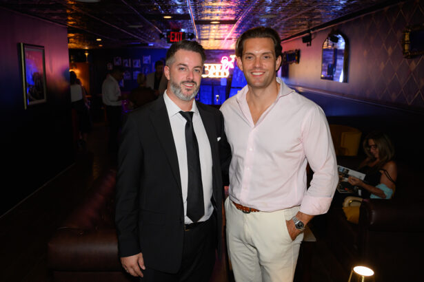 Two men pose together in a dimly lit lounge; one wears a black suit and tie, the other a white shirt and light pants. Neon signs and people are visible in the background. Lifestyle