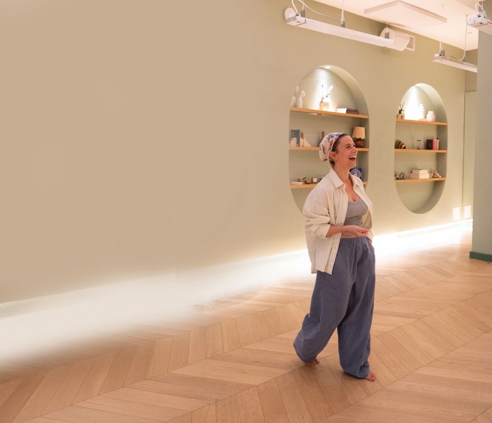 A person wearing loose blue pants, a white shirt, and a headscarf stands barefoot on a wooden floor in a softly lit room with shelves and decorative items on the wall, evoking a sense of movement and quiet connection within the community. Lifestyle