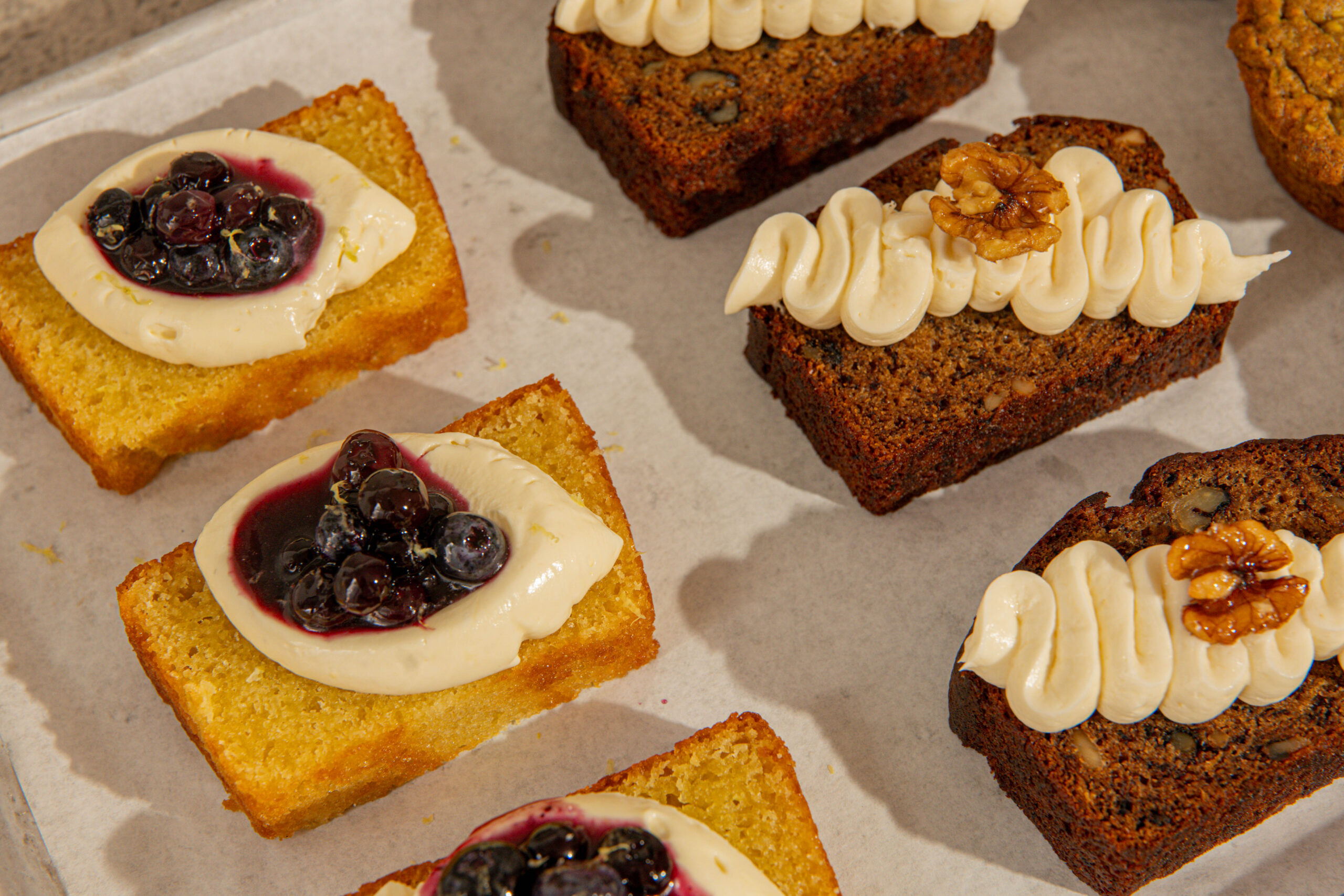 Rectangular mini cakes on parchment; some topped with cream and blueberries, others with piped frosting and walnut halves. These moist, golden-brown cakes—displayed in rows on a baking tray—are perfect for sharing at your next coffee date. Lifestyle