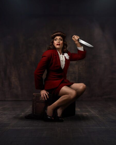 A person in a red suit kneels on a suitcase, holding a large knife overhead. They have styled hair and dramatic makeup, with a theatrical, intense expression against a dark background—capturing the bold spirit of Island City Stage’s 15th Anniversary Season. Lifestyle