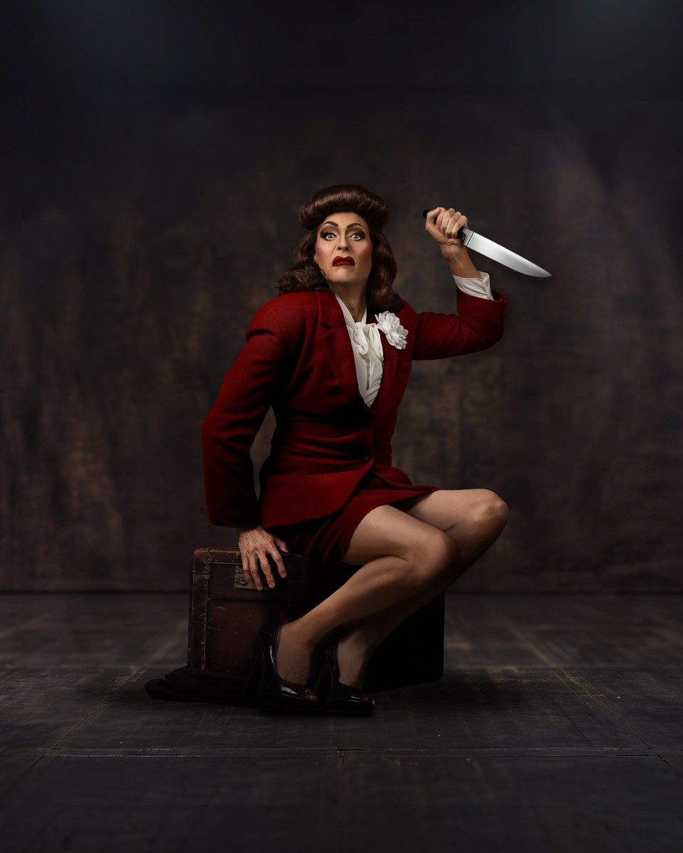 A person in a red suit kneels on a suitcase, holding a large knife overhead. They have styled hair and dramatic makeup, with a theatrical, intense expression against a dark background—capturing the bold spirit of Island City Stage’s 15th Anniversary Season. Lifestyle