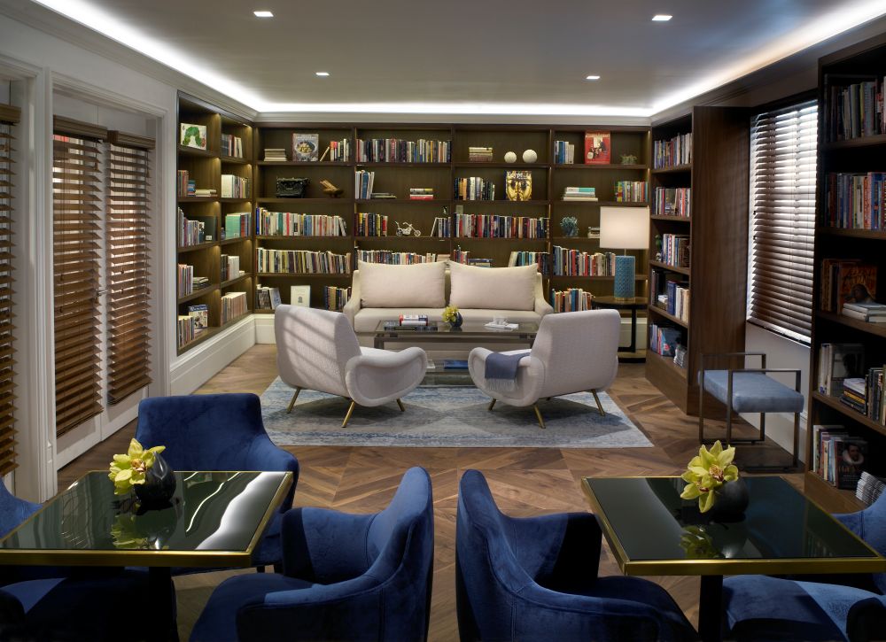 Modern library with bookshelves, a cream sofa, two armchairs, a coffee table, and blue velvet chairs around tables with yellow flowers. Warm lighting and wooden floors create a cozy, inviting atmosphere—perfect for a smart getaway or relaxing afternoon at the Betsy Hotel. Lifestyle