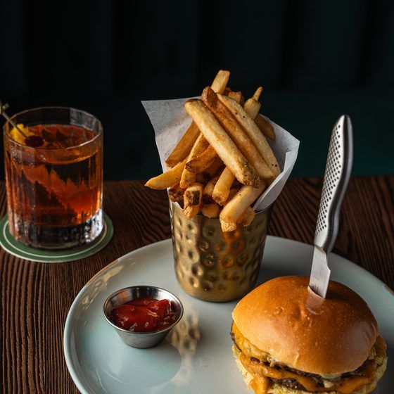 A cheeseburger with a knife in the bun, French fries in a metal cup, a small dish of ketchup, and a glass of expertly mixed cocktail on a wooden table—perfect for savoring in a cozy speakeasy atmosphere. Lifestyle