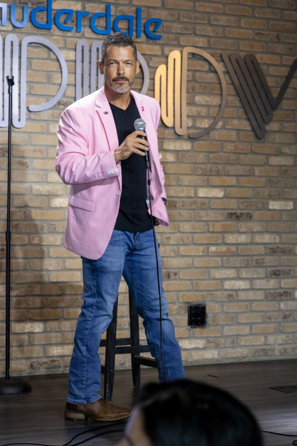 A man in a stylish pink jacket and black shirt stands on a stage holding a microphone in front of a brick wall. Lifestyle
