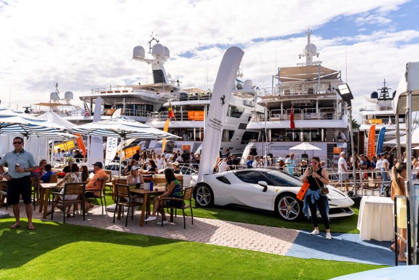 People relax at outdoor tables near a white sports car and luxury yachts docked at a Fort Lauderdale marina. The scene, with banners and umbrellas, goes overboard in showcasing a casual yet upscale event. Lifestyle