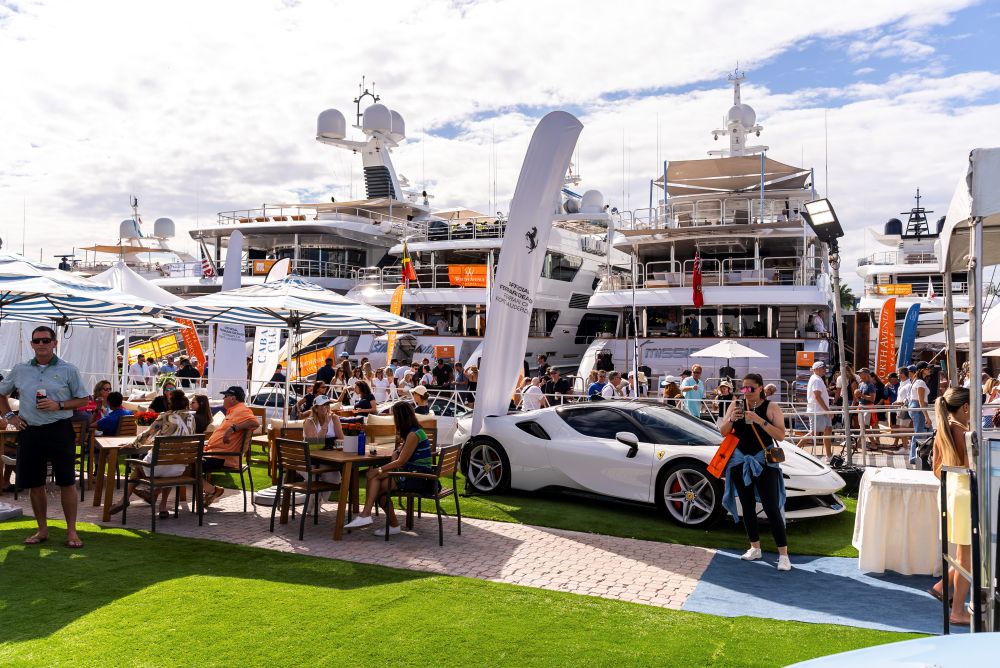 People relax at outdoor tables near a white sports car and luxury yachts docked at a Fort Lauderdale marina. The scene, with banners and umbrellas, goes overboard in showcasing a casual yet upscale event. Lifestyle