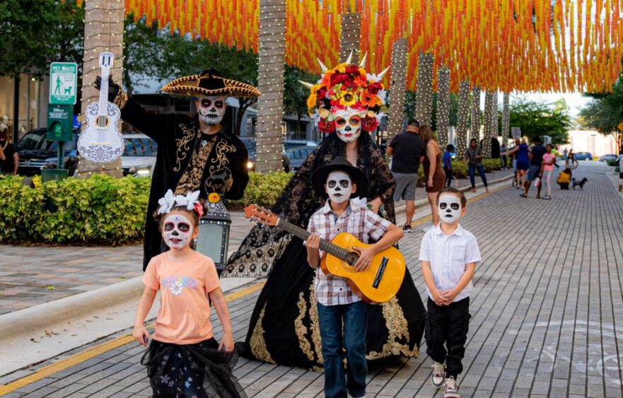 Two adults and two children with sugar skull face paint walk through a CityPlace Doral street decorated with marigolds, wearing traditional Día de los Muertos attire while the children carry a guitar and ukulele. Lifestyle