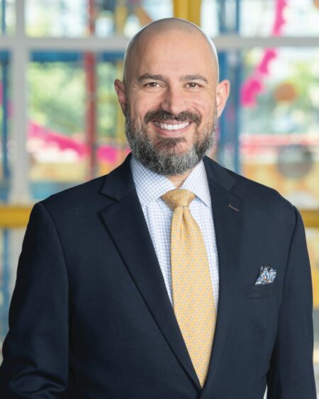 A smiling man with a bald head and beard wearing a dark suit, light shirt, yellow patterned tie, and pocket square stands before colorful windows—Joe Cox looks ahead to building MODS for the next 50 years. Lifestyle
