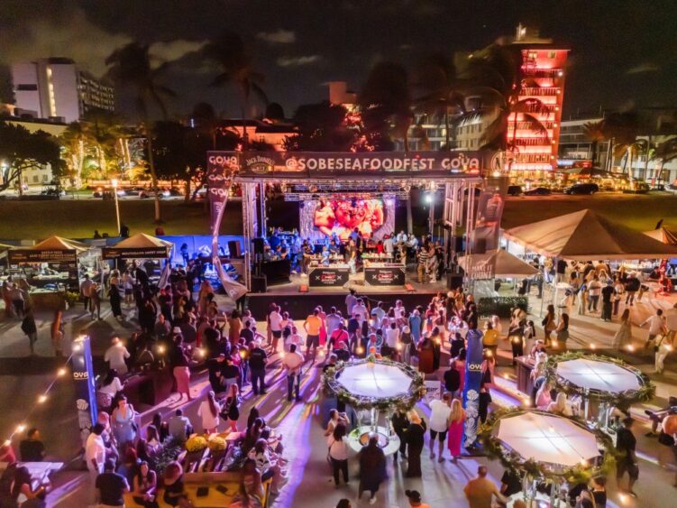 Nighttime aerial view of the South Beach Seafood Festival with a large stage, food vendor tents, and a crowd gathering in front of a giant video screen. Lifestyle