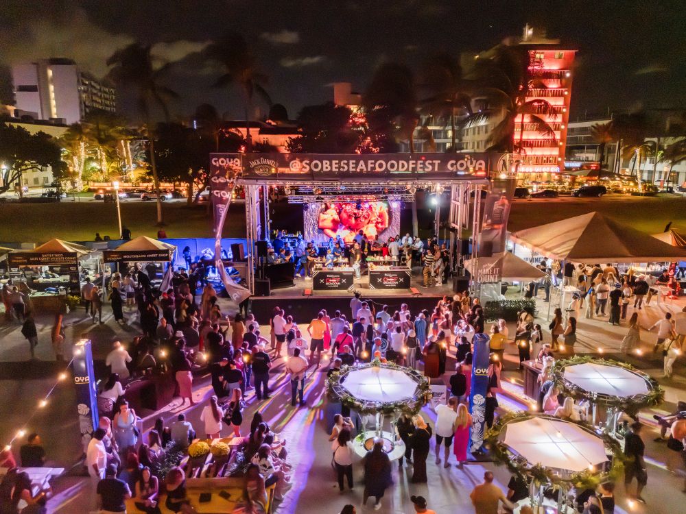 Nighttime aerial view of the South Beach Seafood Festival with a large stage, food vendor tents, and a crowd gathering in front of a giant video screen. Lifestyle