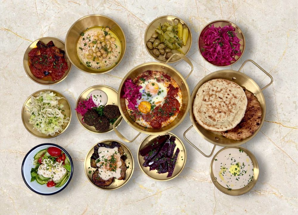 A high-angle view of a Mediterranean feast at Happea’s Mediterranean Grill featuring shakshuka surrounded by hummus, falafel, pickled vegetables, slaw, beets, bread, and salads on a light marble surface. Lifestyle