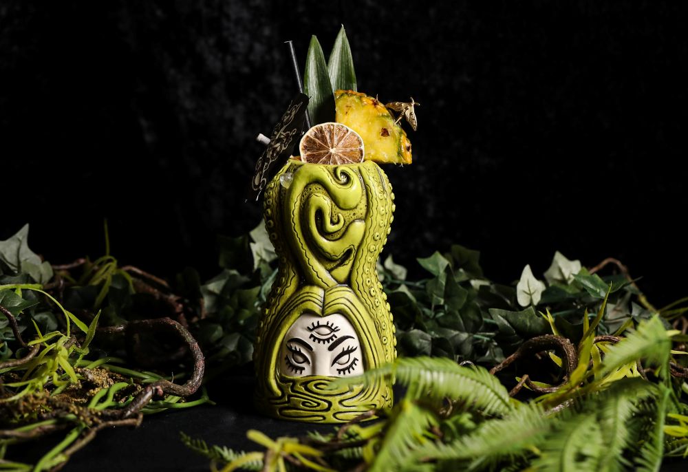A Black Lagoon themed cocktail served in a green ceramic mug shaped like an octopus, garnished with pineapple, a dried lime wheel, and a small umbrella. Lifestyle