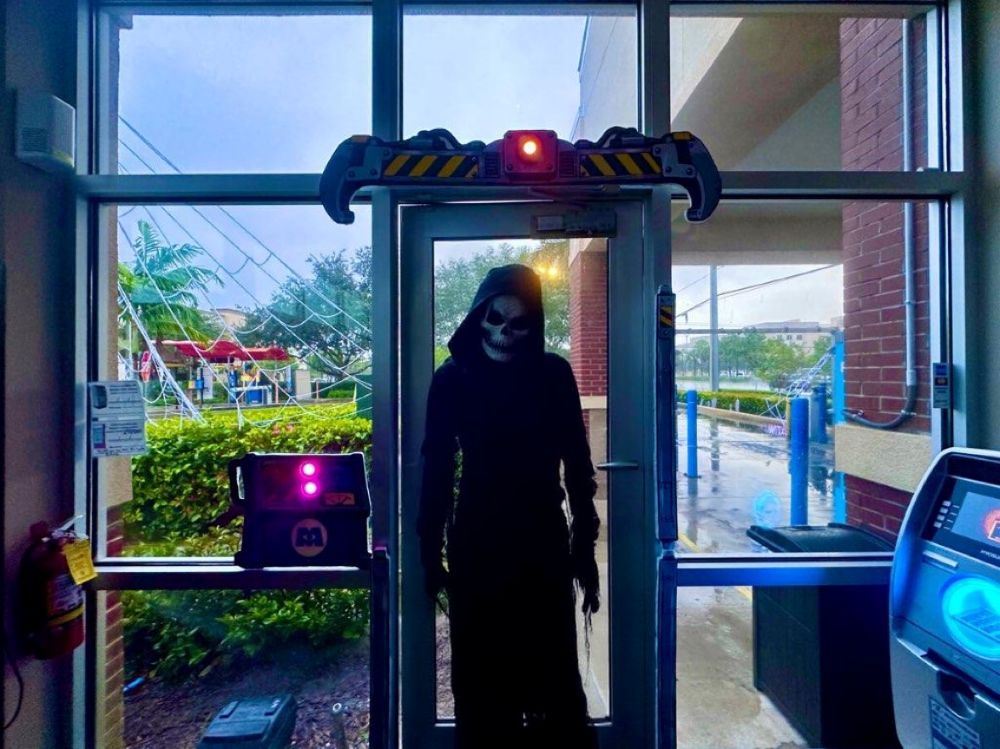 A person in a grim reaper costume stands in the doorway of a haunted car wash with a glowing hazard-striped beam above the entrance. Lifestyle